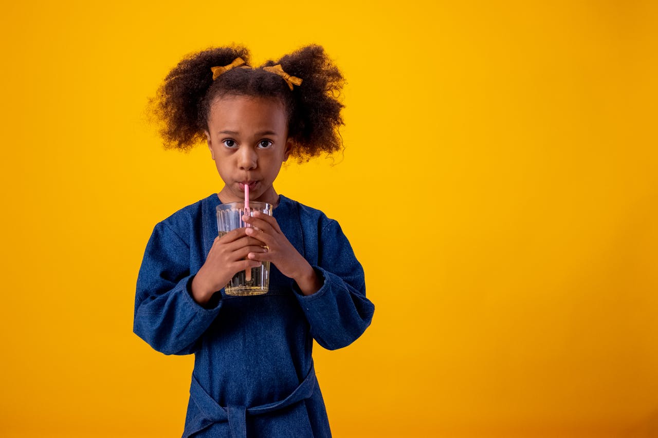 A cute child in a blue dress drinks juice with a straw on a vibrant yellow backdrop.