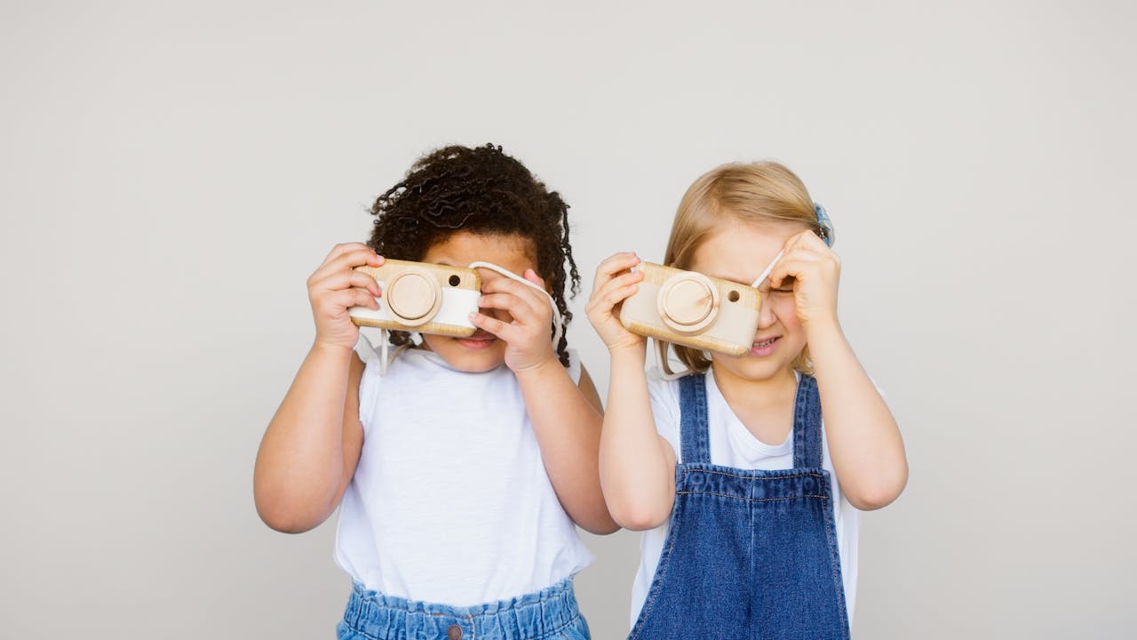 Two children playing with wooden toy cameras in a playful indoor setting, captured in a bright moment.