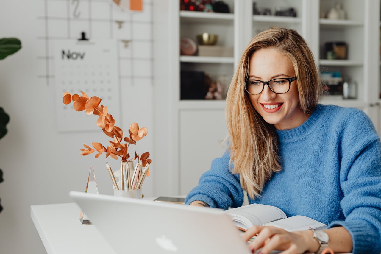 A woman in a bright sweater smiles while working on her laptop at home.