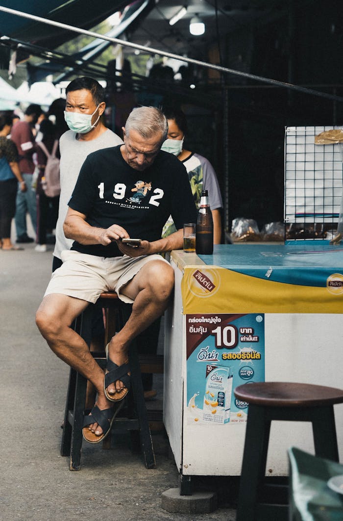 Older man sitting at a market stall checking his smartphone.