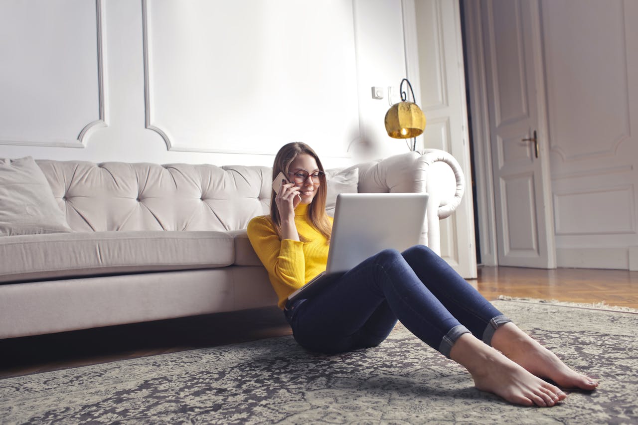 Smiling barefoot female in glasses and casual clothes using laptop and having phone call while sitting on floor leaning on sofa and working on laptop against luxury interior of light living room