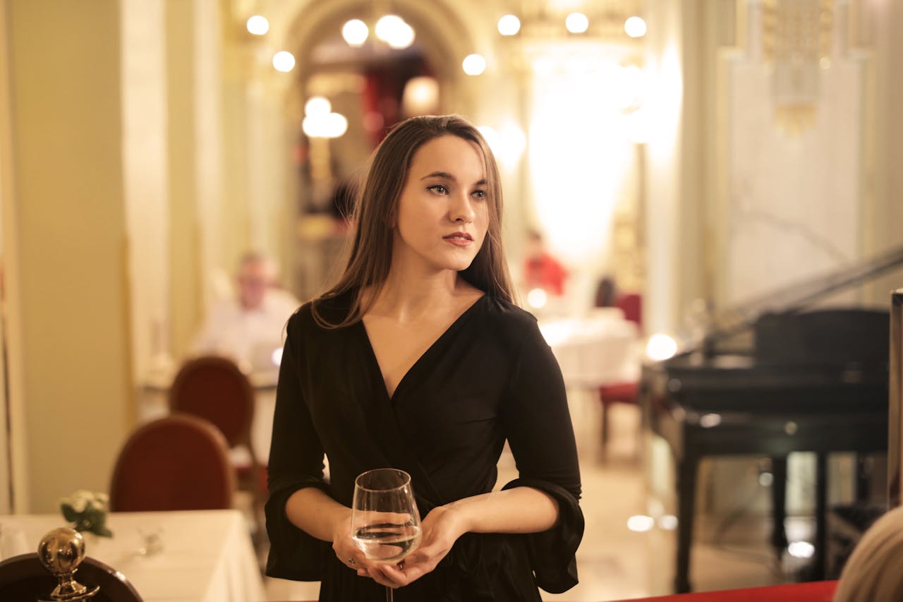 A woman in an elegant black dress holds a wine glass in a luxurious restaurant setting.