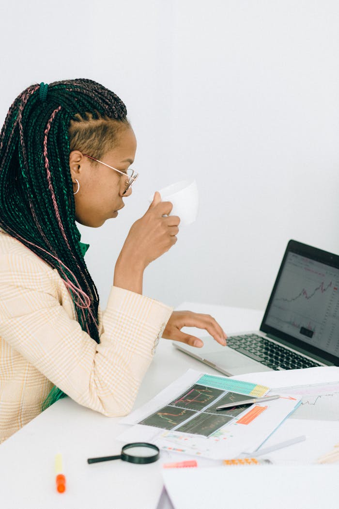A businesswoman with braided hair analyzing stock charts on a laptop, holding a cup.