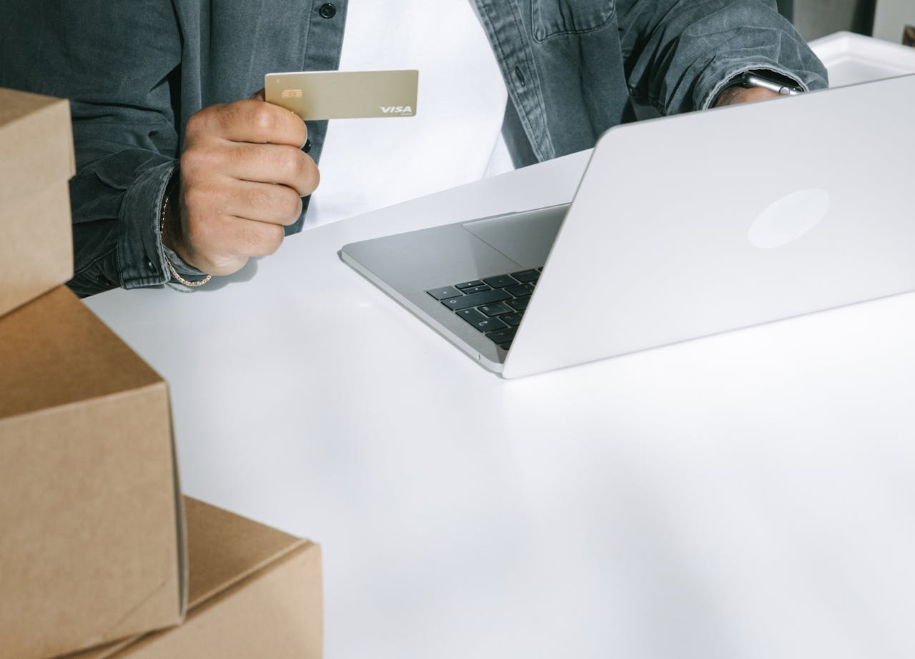 Person using a credit card for online shopping on a laptop surrounded by shipping boxes.