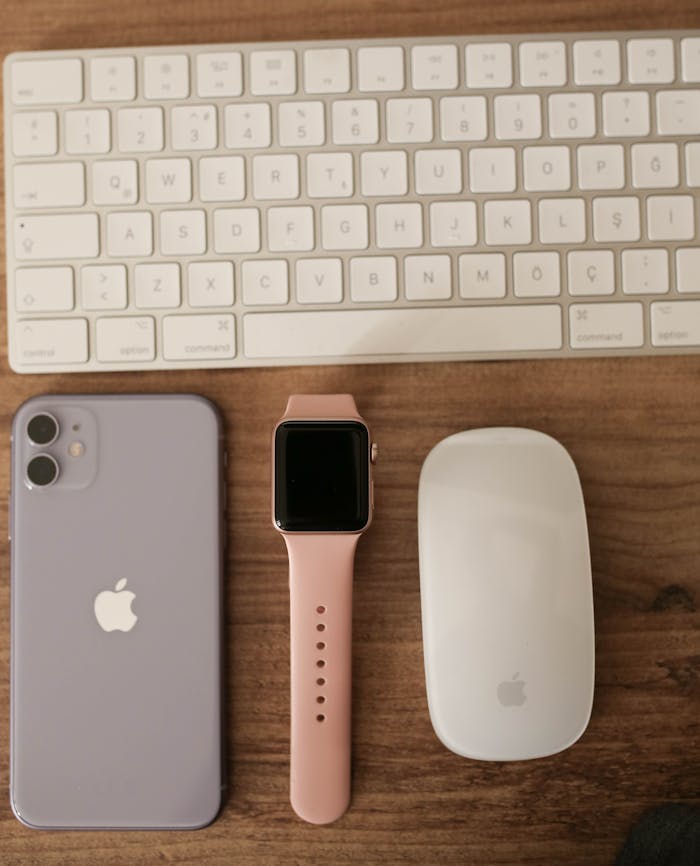 Flat lay of Apple devices including keyboard, mouse, watch, and phone on a wooden desk.