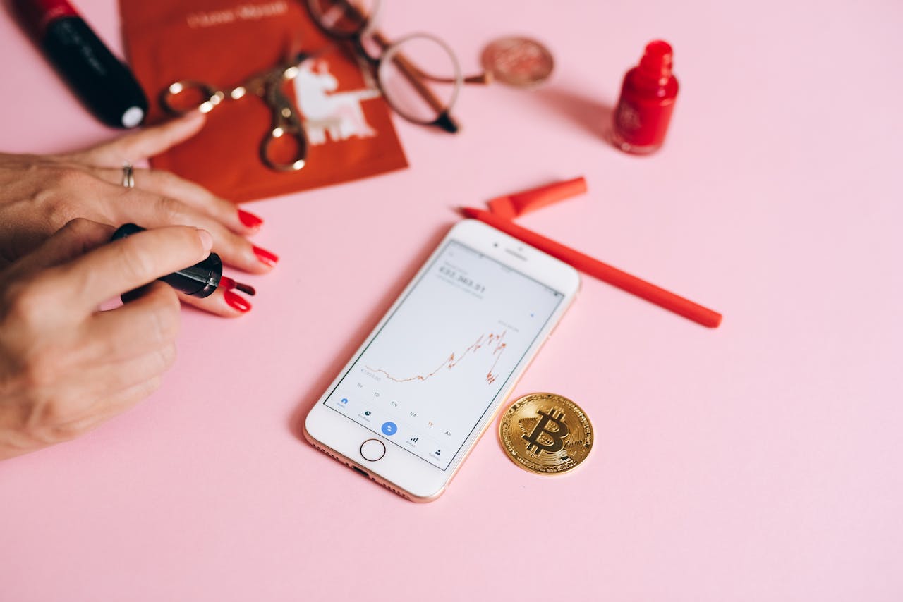 Stylish display of a smartphone with bitcoin, nail polish, and fashion accessories on a pink background.