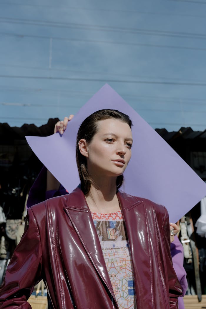 Stylish woman in a maroon leather jacket posing outdoors against a purple backdrop under a clear sky.